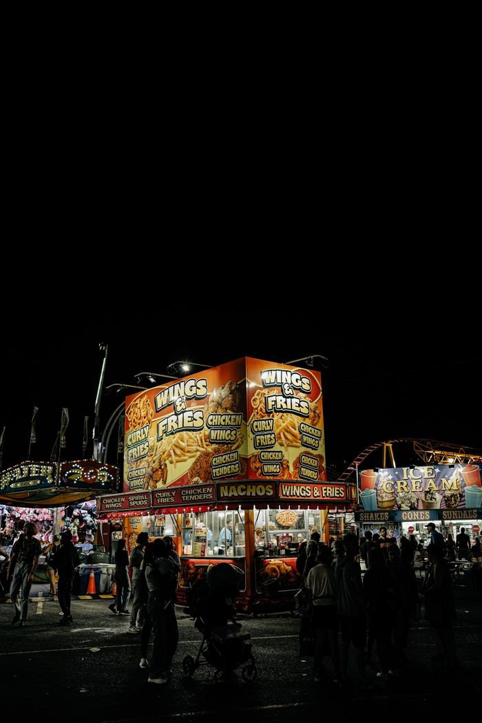 Illuminated food stalls at a night fair serve wings and fries, capturing urban festivity.